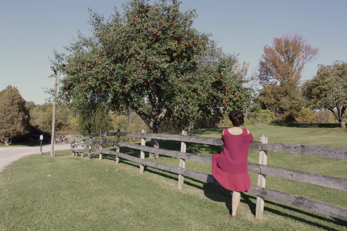 Woman in red dress standing with back to camera in apple orchard